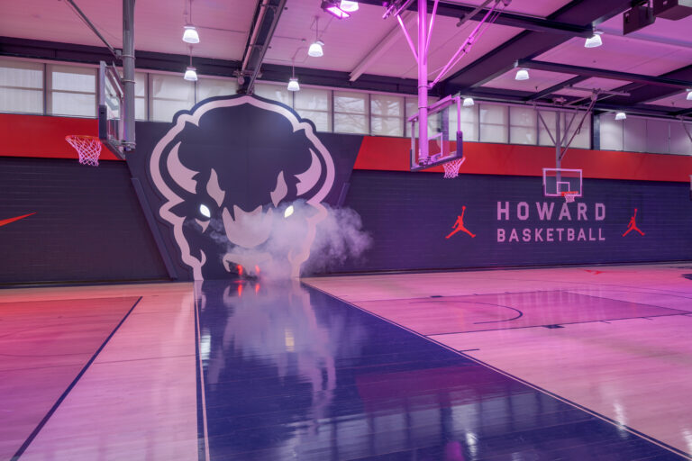 A basketball court with moody red lighting. The space features a large Bison sculptural logo on the wall, emitting smoke for dramatic effect. "HOWARD BASKETBALL" and Jordan brand logos are displayed prominently on the walls.