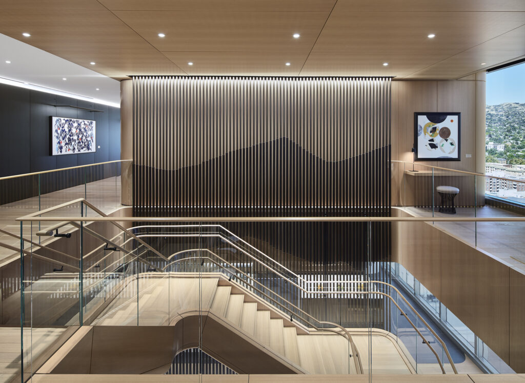 A modern office interior featuring a staircase with glass railings and a large feature wall with vertical metal slats creating a mountain-range pattern.