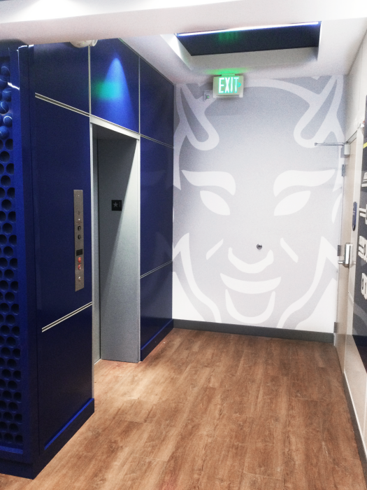 An interior hallway with a blue wall on the left featuring an elevator and a white wall on the right with a large stylized face design. The floor is wood-toned. A green "EXIT" sign is visible above.