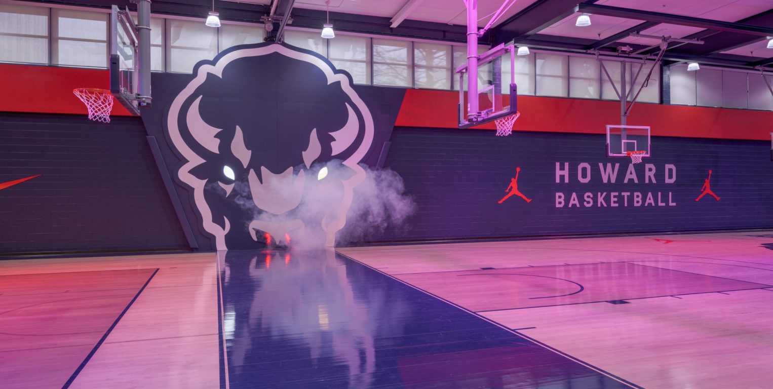 A basketball court with moody red lighting. The space features a large Bison sculptural logo on the wall, emitting smoke for dramatic effect. "HOWARD BASKETBALL" and Jordan brand logos are displayed prominently on the walls.