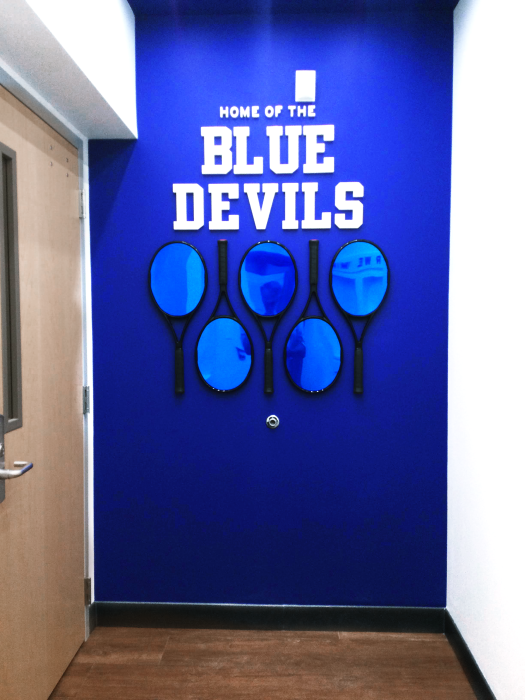 A bright blue wall with white text reading "HOME OF THE BLUE DEVILS". Below the text are five blue-tinted tennis racket shapes arranged in a decorative pattern. A wooden door is visible to the left.